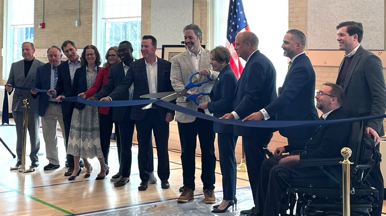 A group of people stands on an indoor stage gathered for a ribbon-cutting ceremony. The person in the middle cuts the ribbon with oversized scissors. 