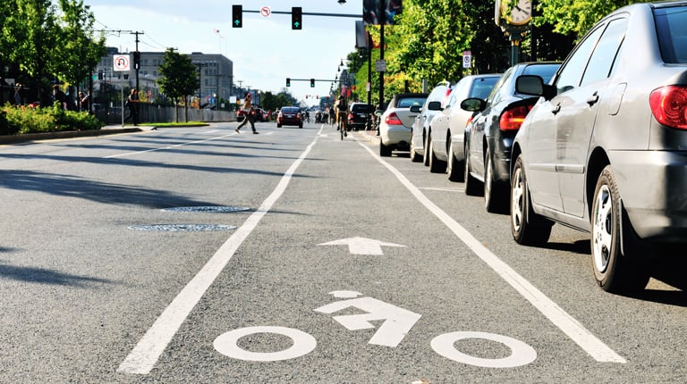Cars parked beside a bike lane, with pedestrians crossing a crosswalk in the background.