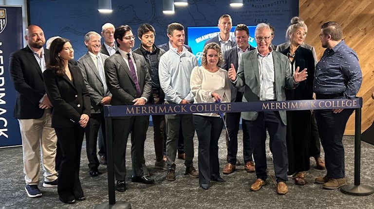 A group of people stands indoors for a photo next to a ribbon labeled "Merrimack College." They are in front of a blue backdrop with a Merrimack College logo.  