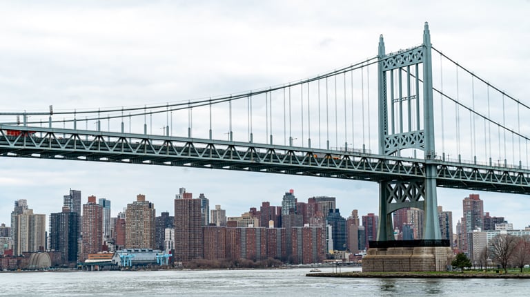 Suspension bridge over water, with the New York City skyline in the background under an overcast sky.