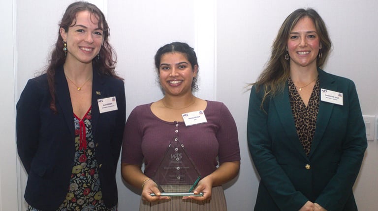Three people stand together and smile at the camera while the woman in the center holds a framed award.