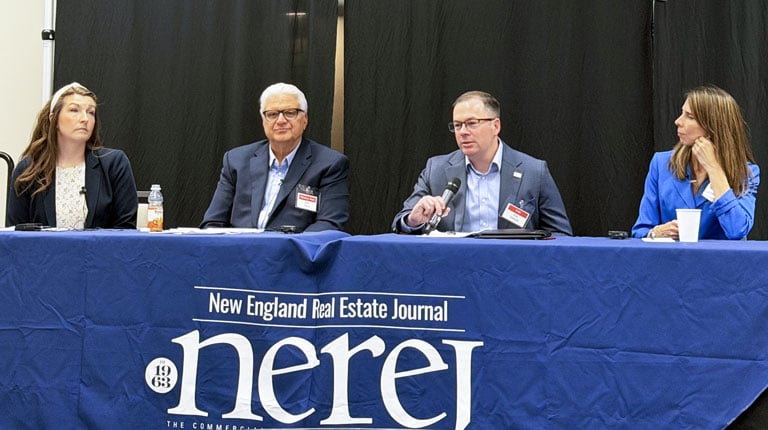 A panel of four individuals with microphones is seated at a table covered with a New England Real Estate Journal tablecloth. 