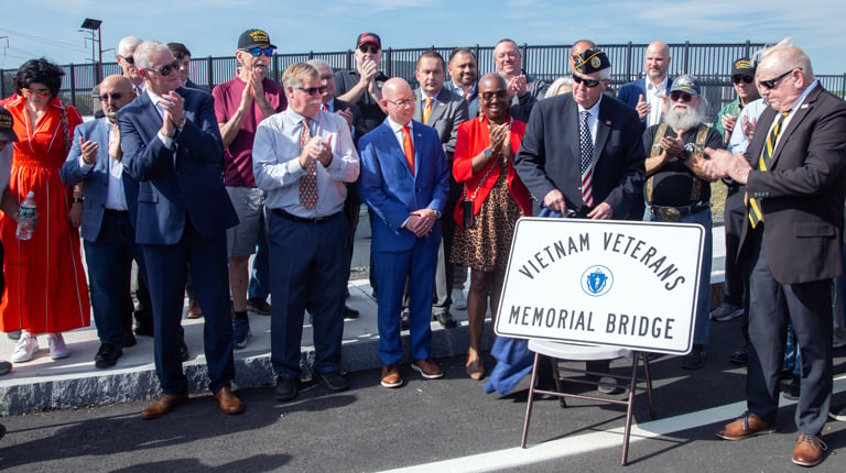 A group of people stand together outdoors, some clapping, around a sign that reads "Vietnam Veterans Memorial Bridge." Several individuals wear military hats.