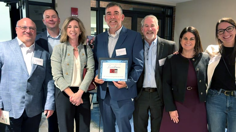 A group of seven people standing together indoors, smiling. One person in the center is holding a certificate. 