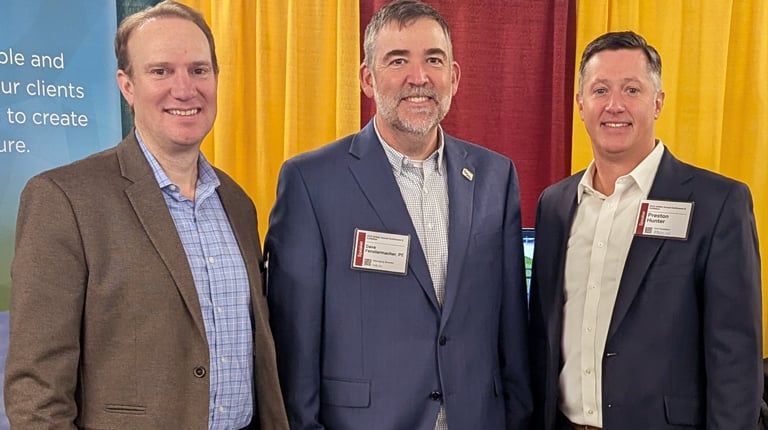 Three individuals standing in front of a banner for VHB, Inc. at a trade show booth. The banner text promotes sustainability and partnership.