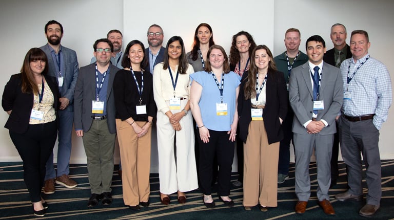 A group of 13 people all wearing business attire with conference badges around their necks. 