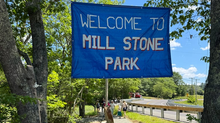 Welcome to Mill Stone Park” banner hanging over a shaded gravel path near the road and trees.