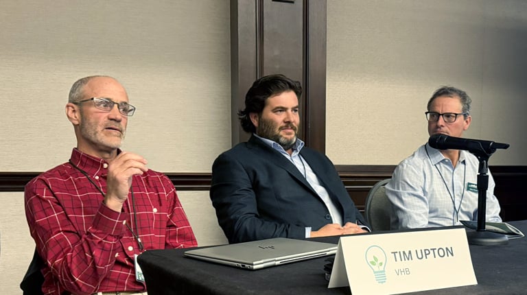 A panel of three individuals seated at a table with nameplates, speaking at an indoor event. 