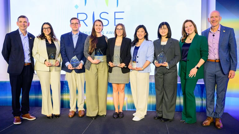 Chief Operating Officer Tom Jackmin, Sara Yasin, Connor Burke, Marisa Blackwood, Madi Karis, Susan Lee, Hyeajin Kim, Chief People Officer Keri Kocur, and President Bill Ashworth stand on stage. The six award recipients in the center hold glass trophies.