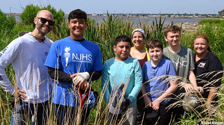 Six students and community volunteers stand in tall grass under a clear blue sky.
