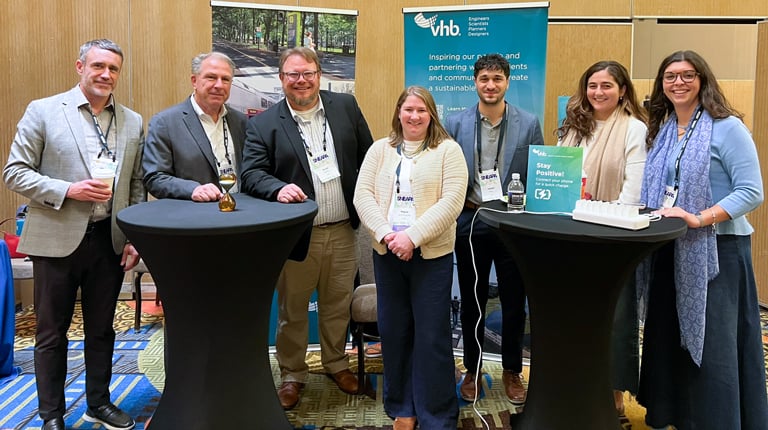 A group of seven people stands together indoors, smiling at a conference booth. The booth has a banner displaying the VHB company logo and information.  