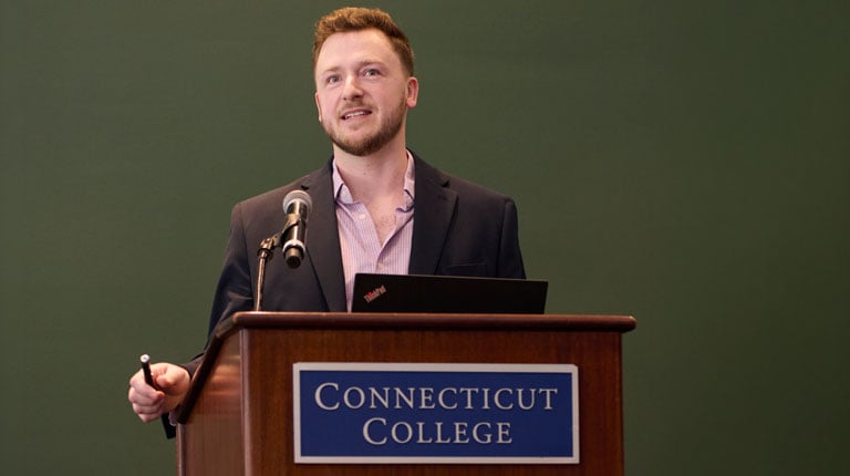 A person stands at a podium with a sign reading "Connecticut College," speaking into a microphone and holding a presentation clicker.