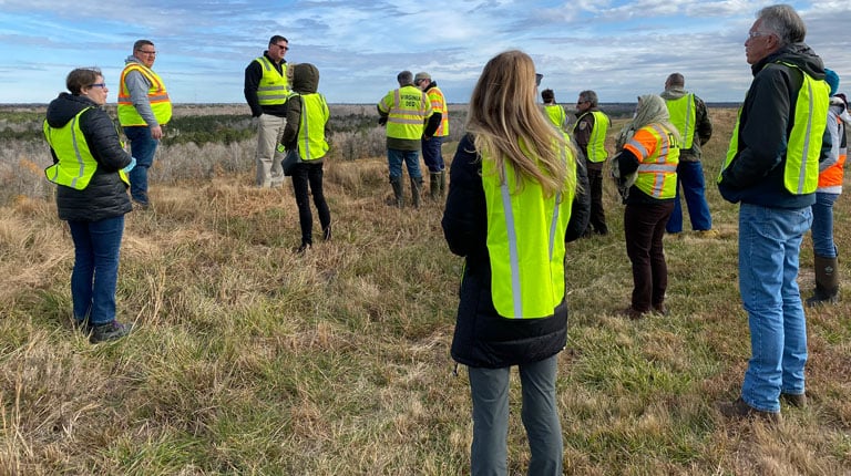 Group of tour participants in safety vests standing on a grassy capped landfill cell overlooking the surrounding landscape.