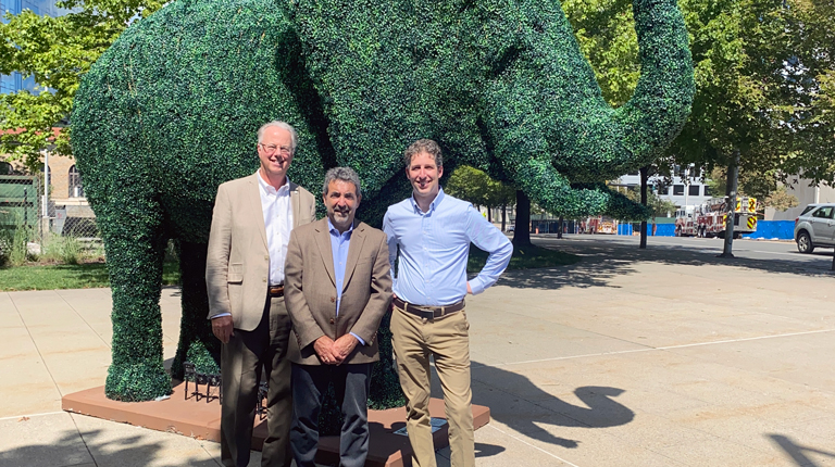 Three men stand in front of a green, elephant topiary on a sidewalk.