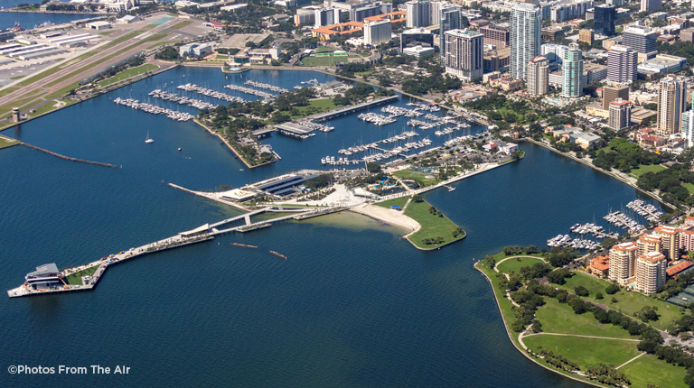 An aerial color photo of the city of St. Petersburg, Florida, shows tall buildings in an urban downtown area located close to Tampa Bay shoreline and the extensive tree canopy of South St. Petersburg peninsula.