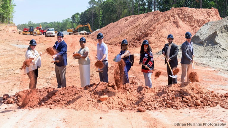 People wearing hard hats standing in line on construction site, holding shovels, tossing soil at groundbreaking.