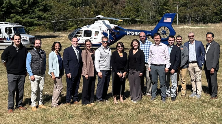 Group of professionals standing in a field with a helicopter and emergency vehicles in the background.