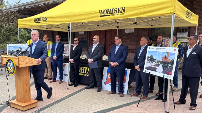 A man speaking at a podium with the Wohlsen Construction banner, surrounded by several individuals standing under a tent.