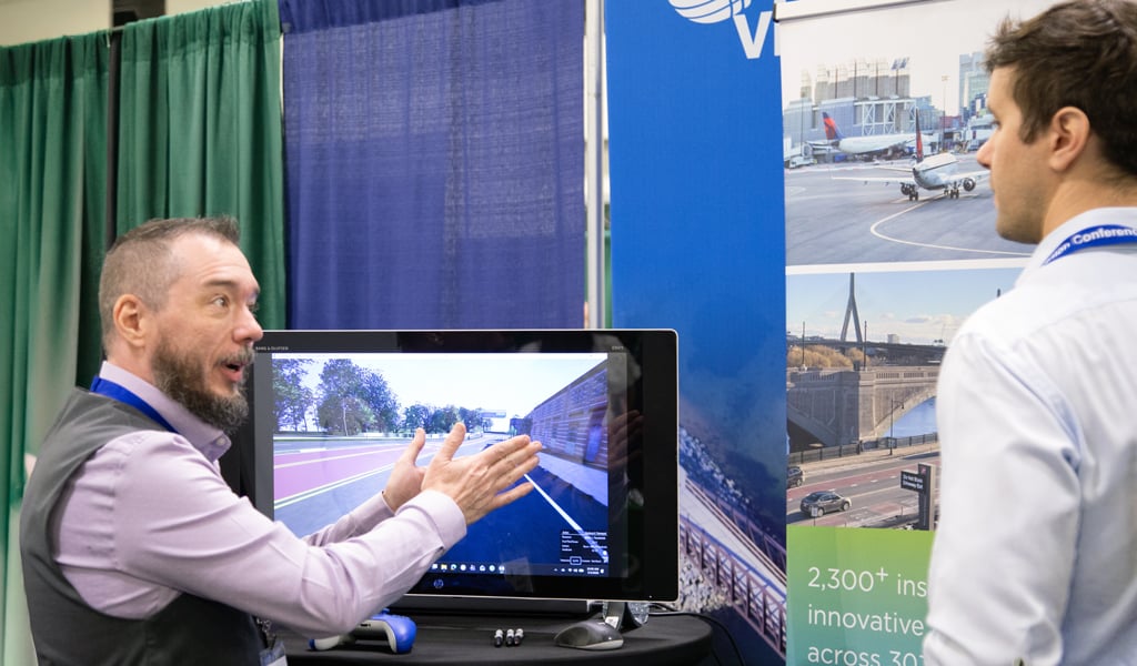 A person is enthusiastically gesturing towards a display screen showing a visualization of an infrastructure project at a trade show booth.  