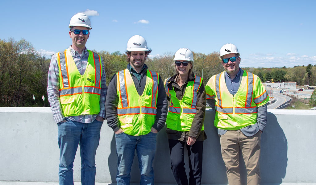 Four people are standing on a paved surface in front of a concrete barrier. They are wearing safety vests and hard hats with the VHB logo.  