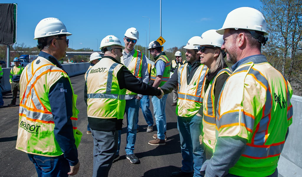 A group of people wearing safety vests and hard hats stand on a road under construction, engaged in conversation. One person is shaking hands with another.  