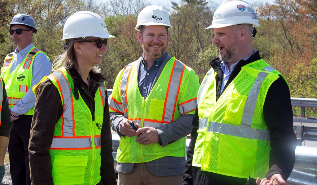 A group of individuals wearing hard hats and high-visibility vests stands on a road near a guardrail. Some have logos on their helmets, including one that reads "VHB."  