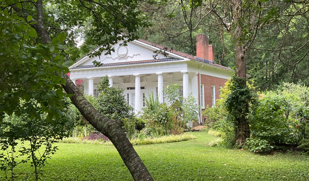 A quaint brick house with white columns is nestled in a lush, green garden surrounded by trees. 