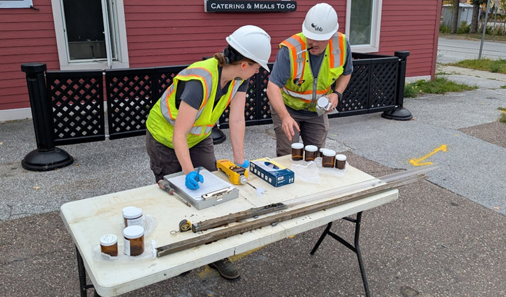 Two construction workers in reflective vests are examining equipment on a table in front of a brick building.