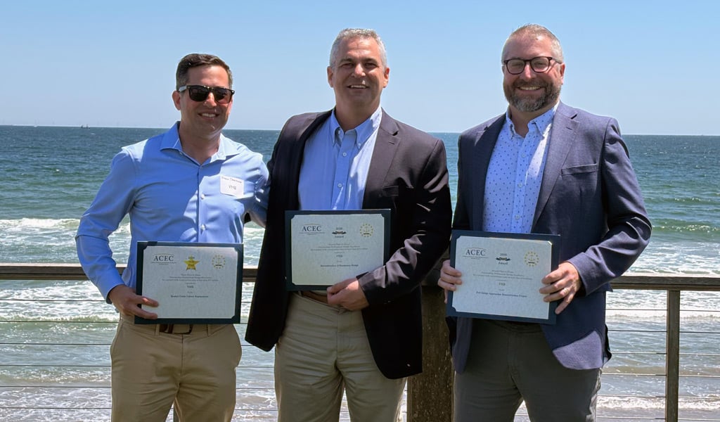 Three suited men with award plaques in front of the beach.