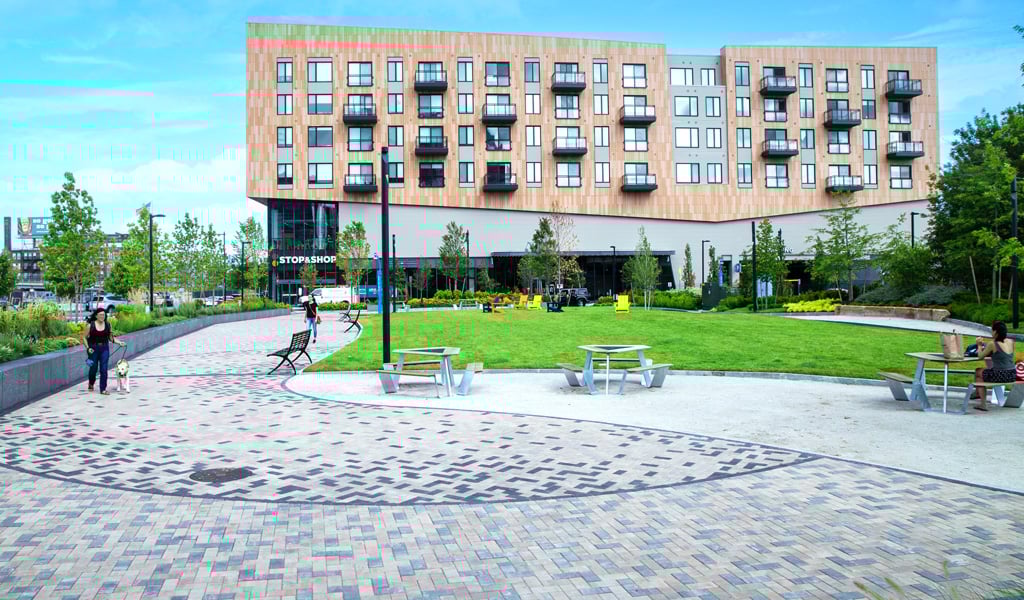 View of a public space with several people and seating areas, a patterned pavement, green lawns, and a large building.