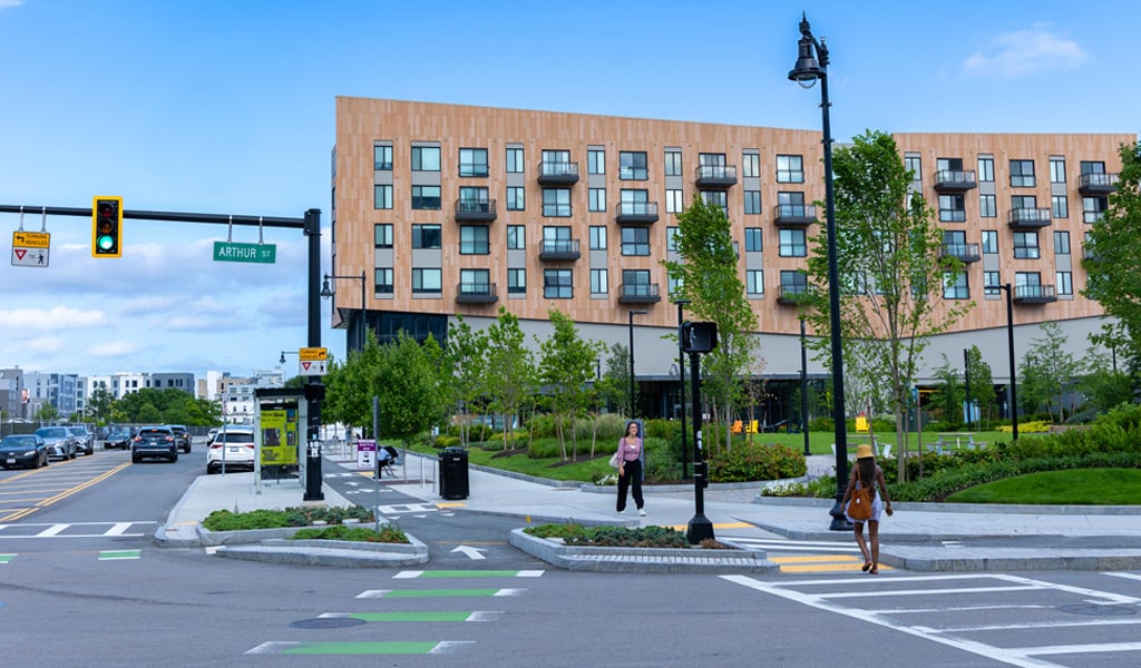 View of a busy intersection in front of the Hyatt Regency hotel on a clear day, with pedestrians crossing, traffic lights, and lush greenery visible.