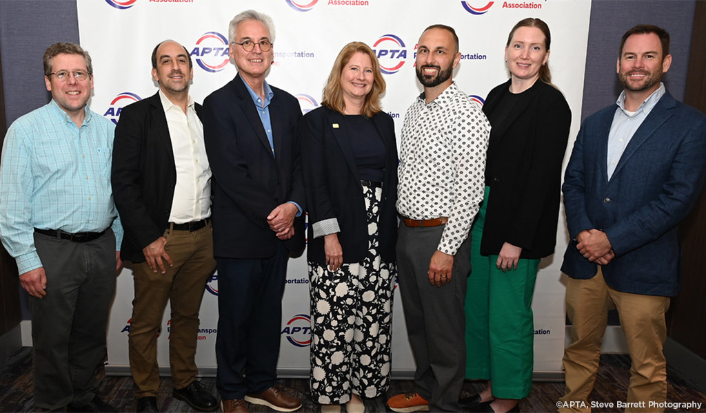 Group of seven professionals standing together at an APTA (American Public Transportation Association) event, smiling for the camera in front of a backdrop with APTA logos.