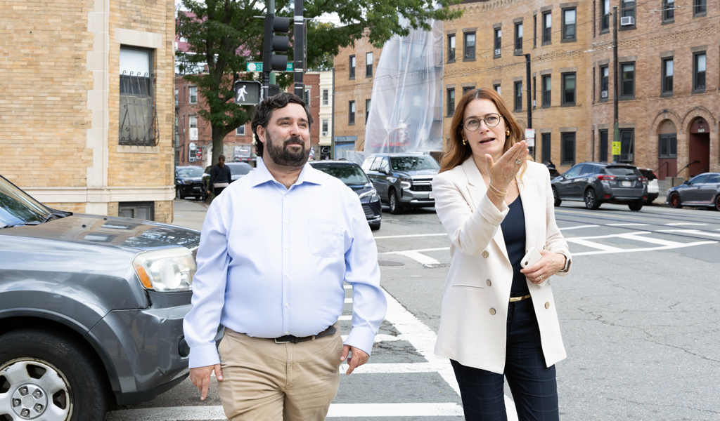 Two adults walking and talking on a city street corner, with brick buildings and a street light in the background. 