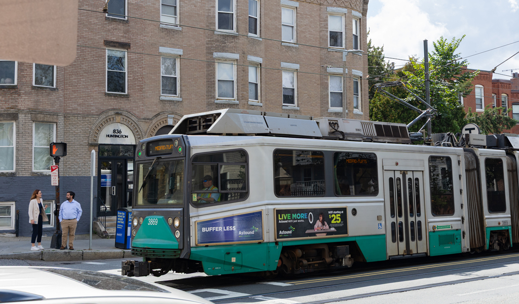 Two people waiting at a tram stop with a green and blue tram passing by in a city street lined with buildings.