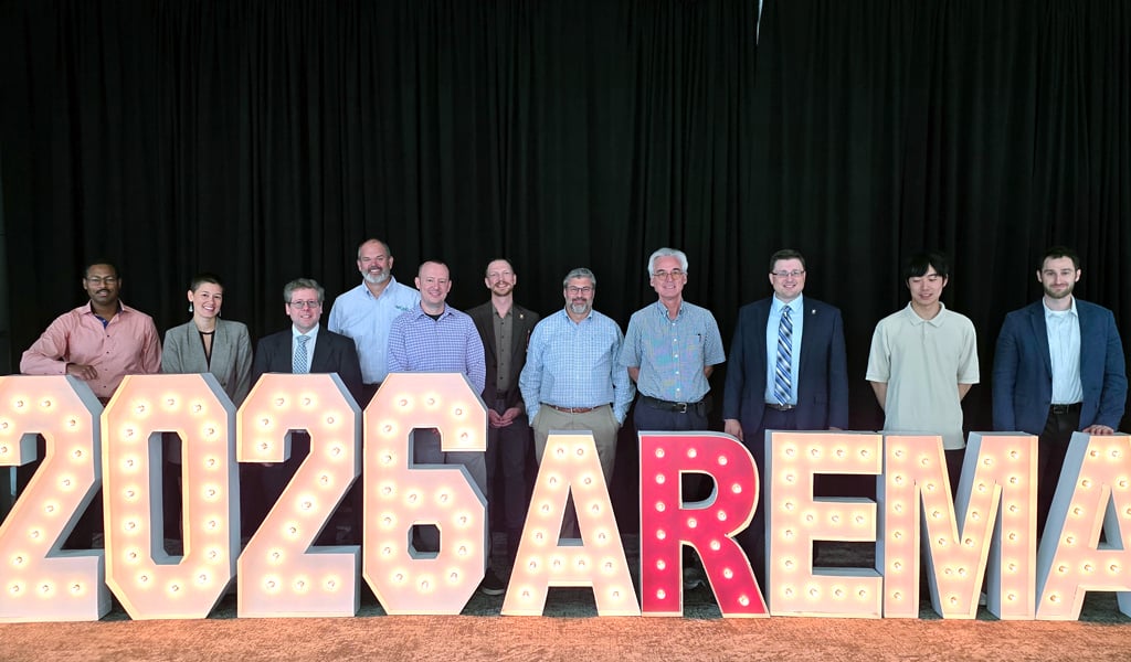 A group photo of 11 VHB team members standing behind a large floor sign that reads 2026 AREMA. 