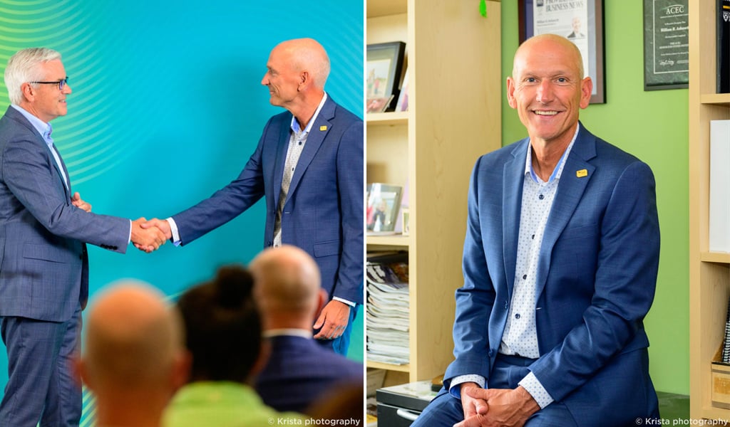 Two men in blue suits on stage in front of a colorful backdrop and man in blue suit sitting smiling.