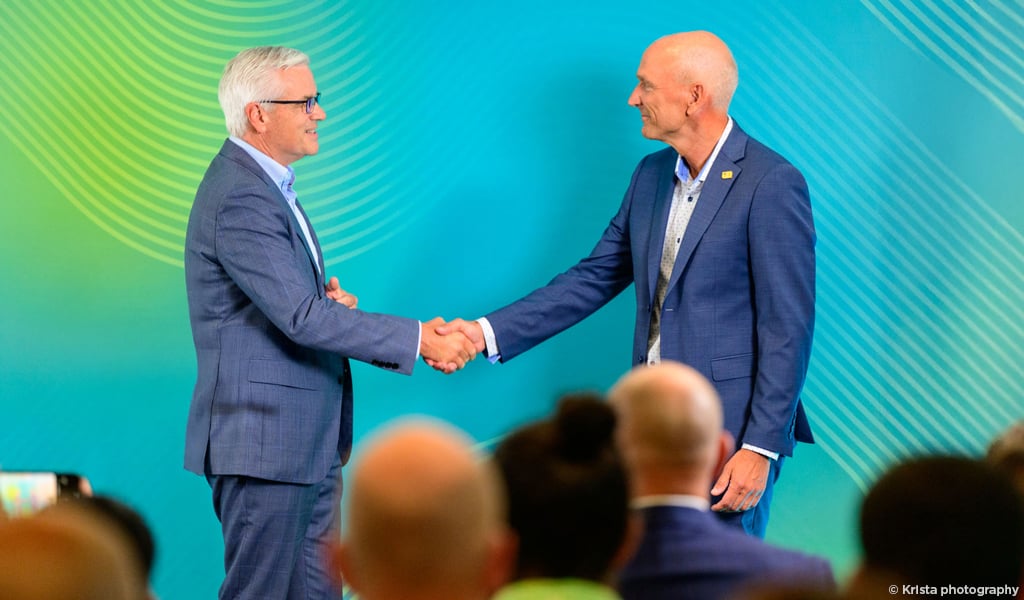 Two men in blue suits on stage in front of a colorful backdrop.