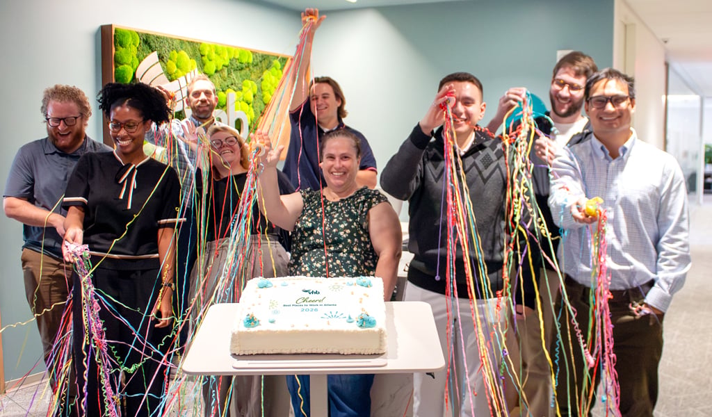Employees stand around a celebration cake in a VHB office while launching confetti streamers.