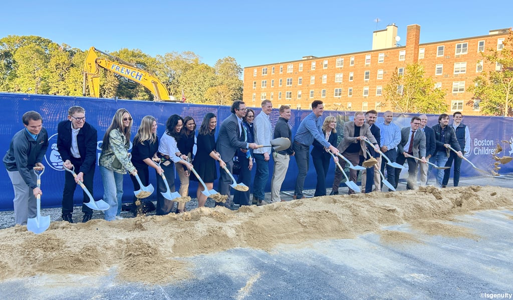 Group breaks ground with shovels at a construction site, with an excavator and Boston Children’s banner behind them.
