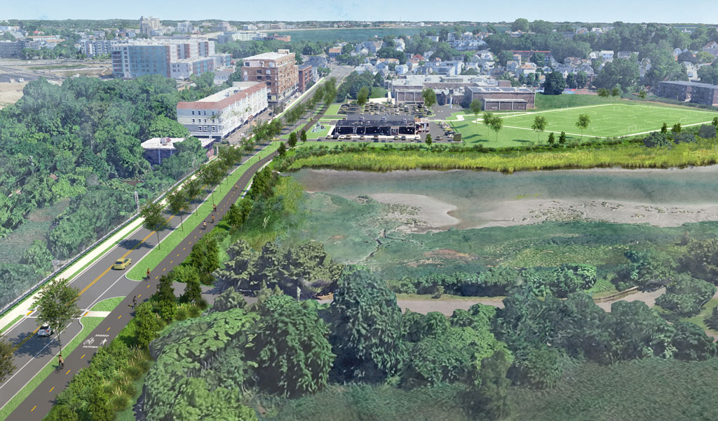An aerial view of a marshy area and a sports field, showcasing a winding road lined with trees, nearby modern buildings, and expansive green spaces.