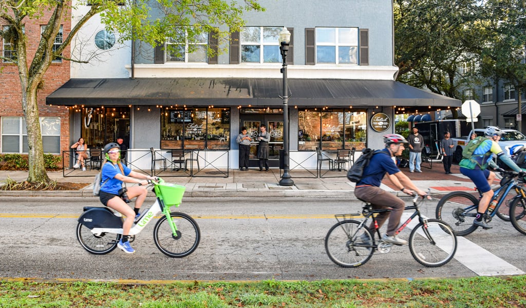 Several cyclists ride along a city street past storefronts and outdoor seating.