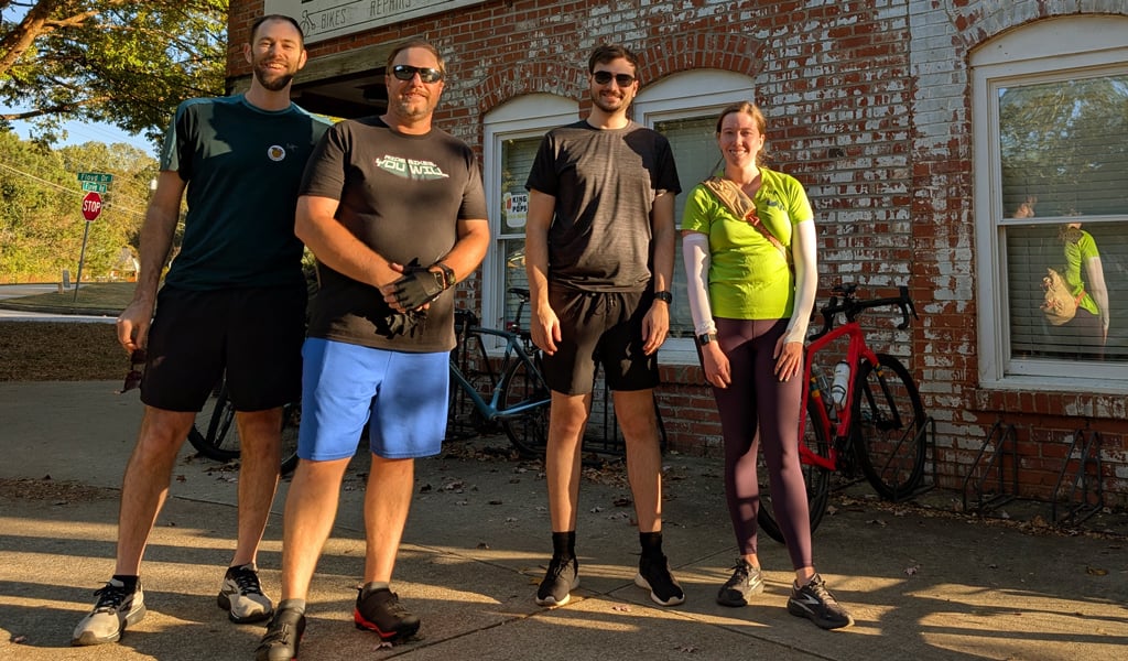 Four smiling VHBers smiling and taking a break from biking in front of a brick building.
