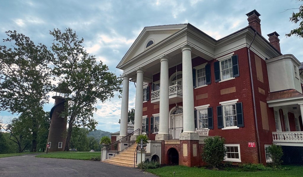 Historic red brick mansion with white columns and black shutters, viewed from the front drive.