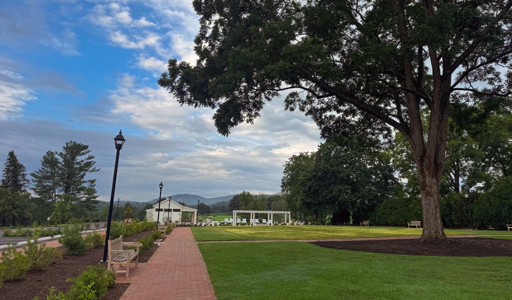 Expansive lawn with a brick pathway, benches, mature shade trees, and event space structures overlooking the surrounding landscape and distant mountains.