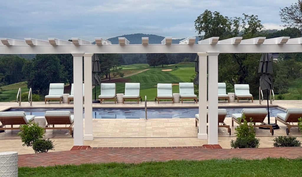 Pool terrace with lounge seating and a white pergola overlooking rolling green fairways, trees, and distant hills under a cloudy sky.