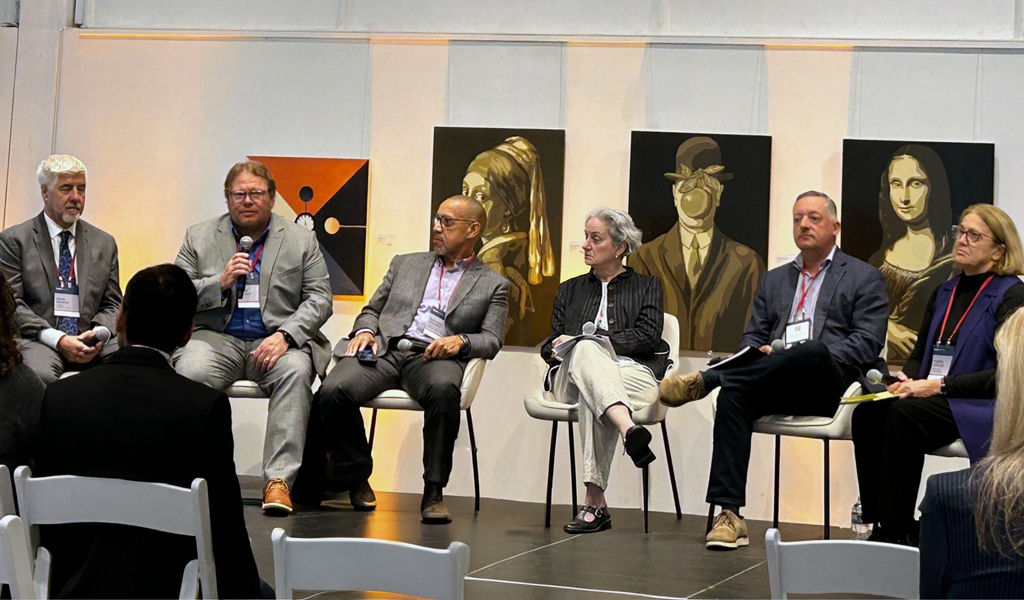Panel of six speakers seated onstage at a conference. The audience is visible in the foreground, listening attentively.