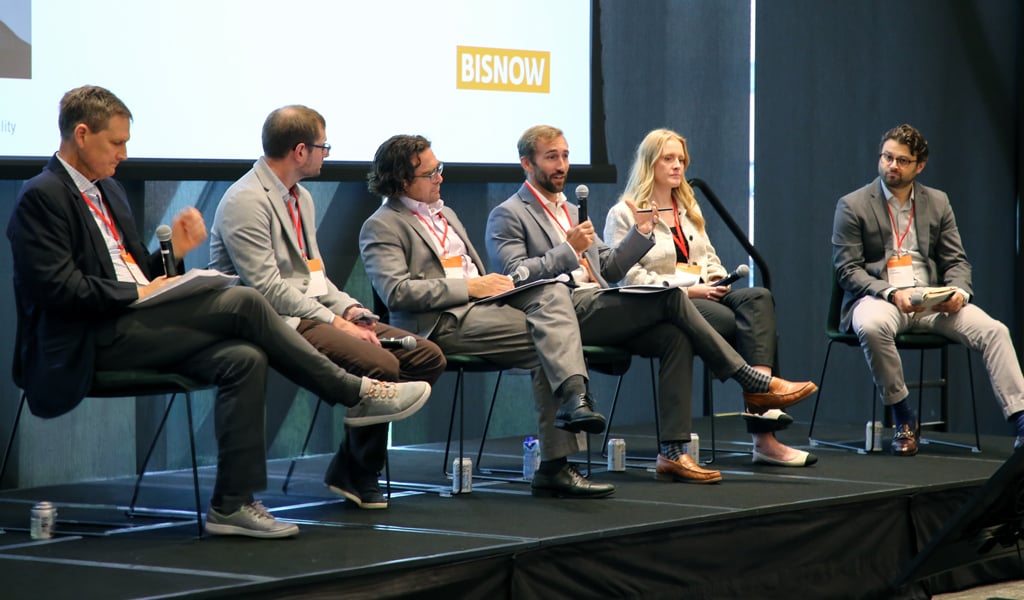 A panel of six individuals seated on stage at a Bisnow event. They are engaged in a discussion, each with a microphone.