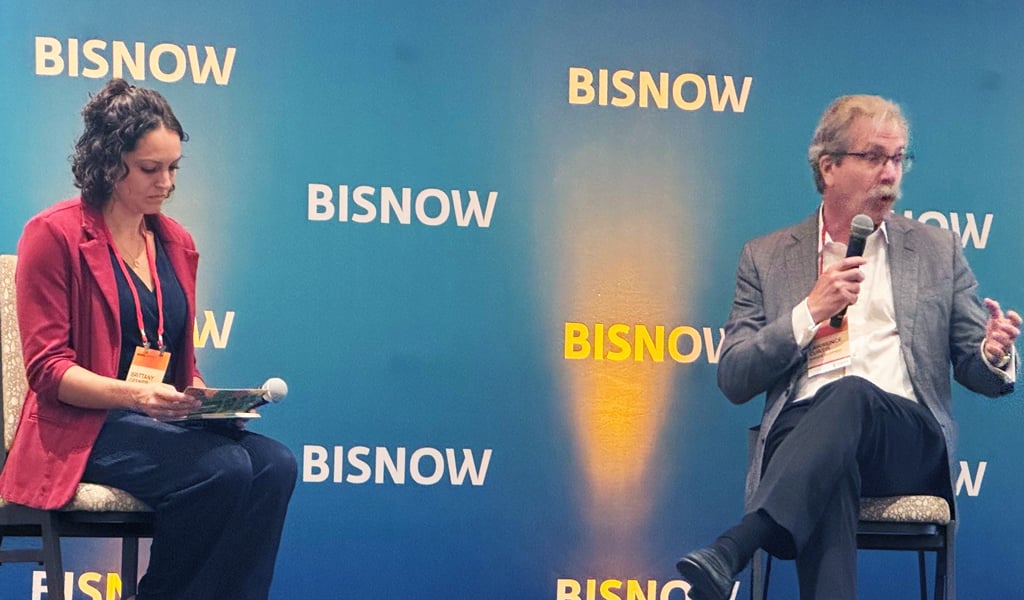 Two individuals participating in a panel discussion at a Bisnow event. One is seated on the left, looking at notes, and the other is holding a microphone, speaking animatedly.