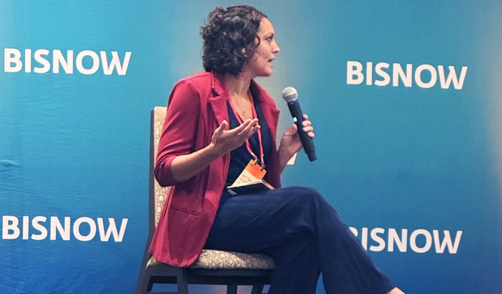 A person in a red blazer and dark pants sitting on a stage, speaking into a handheld microphone at a BISNOW event.