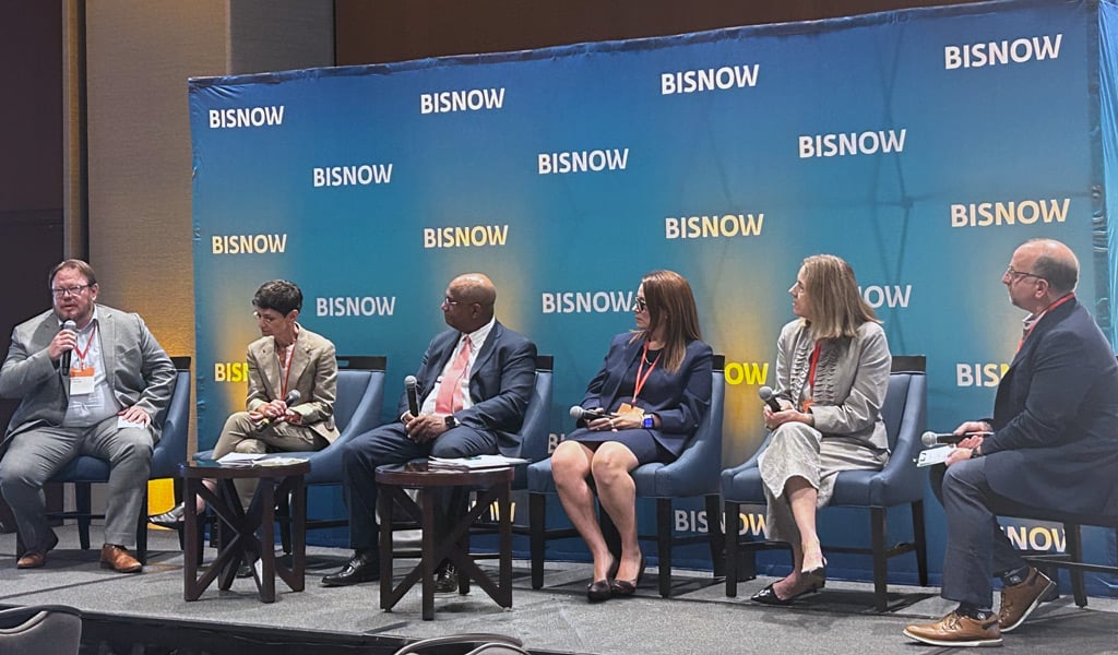 A panel of six individuals seated on a stage with a "Bisnow" backdrop. 